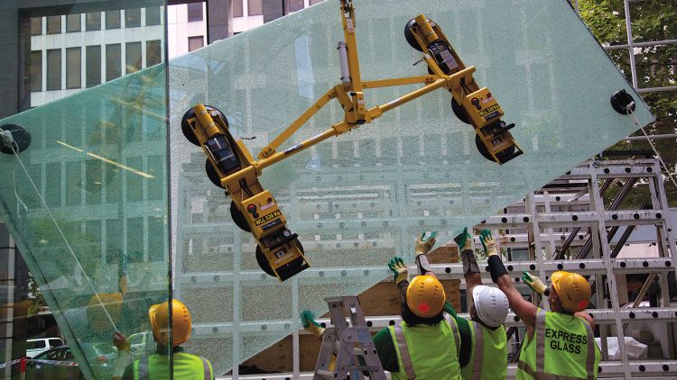 Express Glass workers in safety gear are using a yellow glass-lifting device to install a large glass panel at a building site.