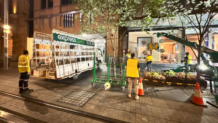 A nighttime street scene with Express Glass emergency response workers in yellow vests near a retail area in George Street Sydney. There are safety cones and a large green trolley, with a retail shop visible in the background.