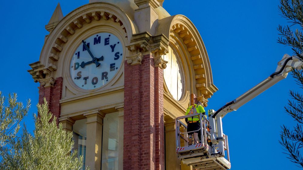 An Express Glass worker on a boom lift is repairing the famous Dimmeys clock on a tower, set against a clear blue sky.