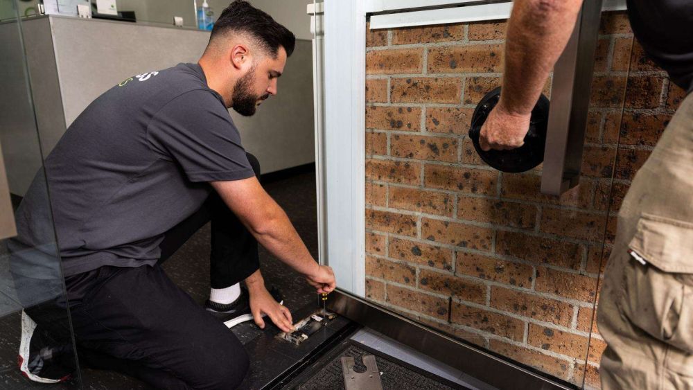 Two Express Glass tradesmen working on repairing a window; one person is kneeling and adjusting a window component, while the other is holding a glass lifting tool.