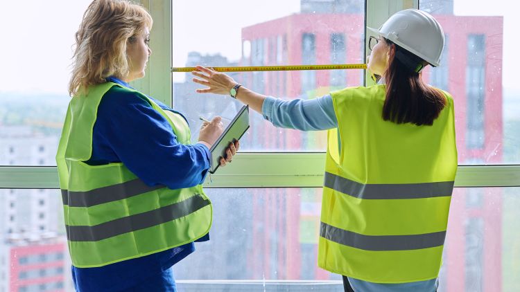 Two women in safety vests are inside a building. One is measuring a window for AS 1288 compliance with a tape measure, while the other is taking notes on a clipboard.