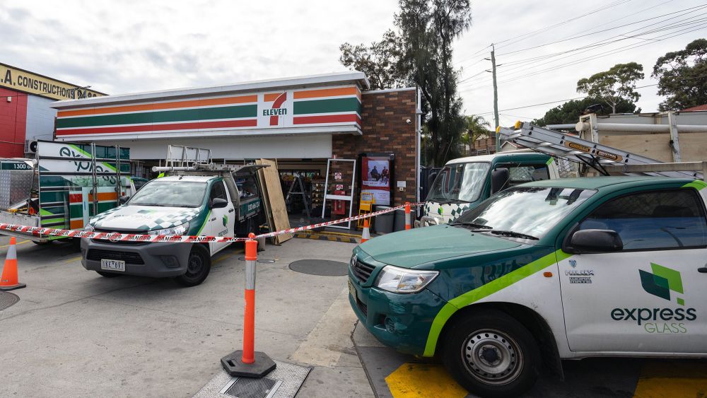 A 7-Eleven convenience store with several Express Glass service trucks outside. The area is marked with traffic cones and caution tape.