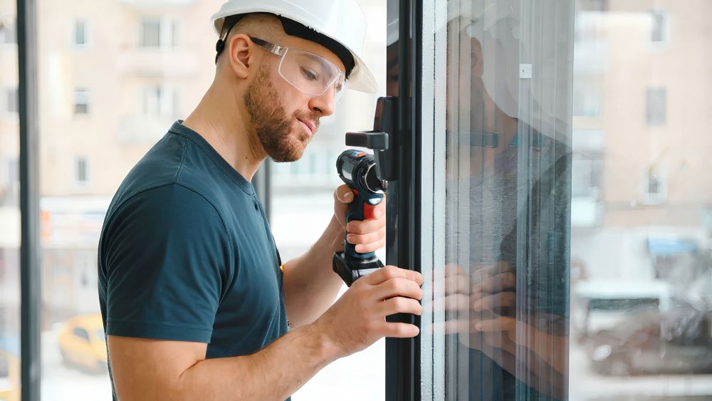 An Express Glass maintenance engineer wearing a hard hat and safety goggles is using a power drill on a window frame in an indoor high rise setting.
