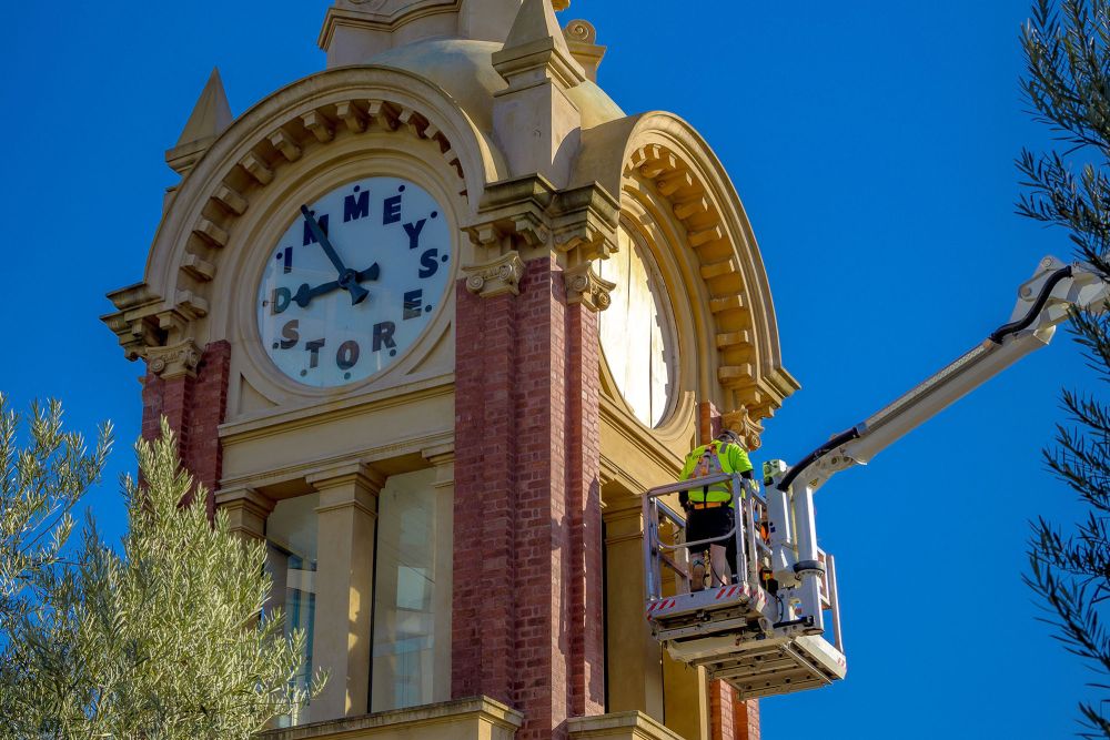 An Express Glass worker on a boom lift is repairing the famous Dimmeys clock on a tower, set against a clear blue sky.