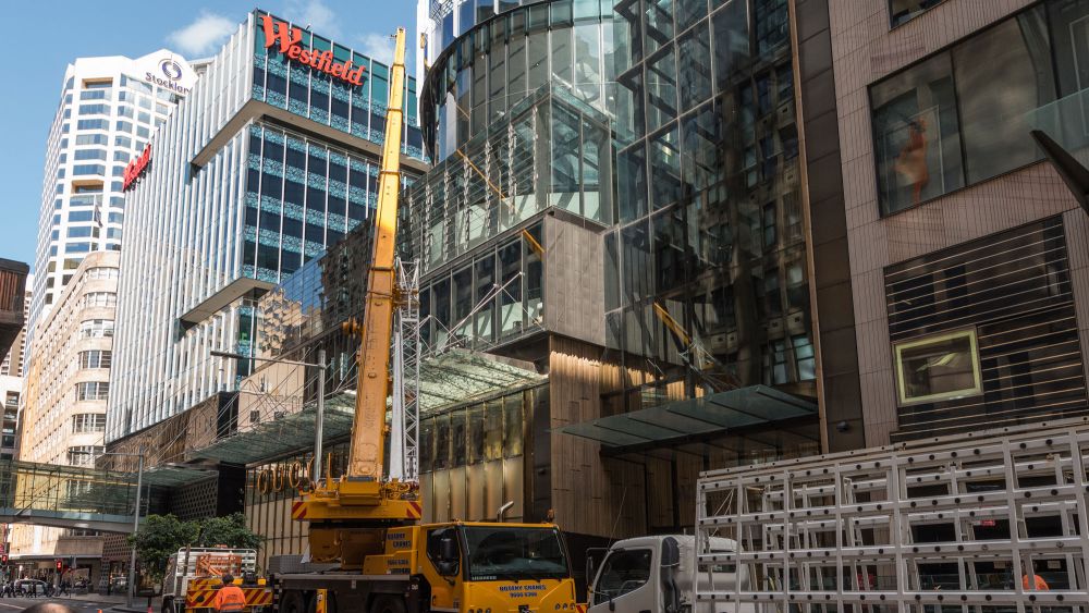 A busy city street with a large yellow construction crane, trucks, and modern glass buildings. The crane is working to lift a large glass pane up to the facade of a commercial building.