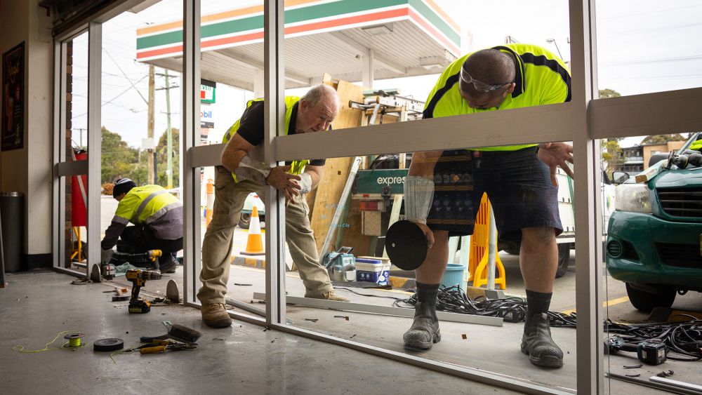 Three Express Glass emergency response workers in high-visibility vests are repairing a 7-Eleven storefront window. Tools and equipment are scattered on the floor, and a 7-Eleven forecourt is visible in the background.